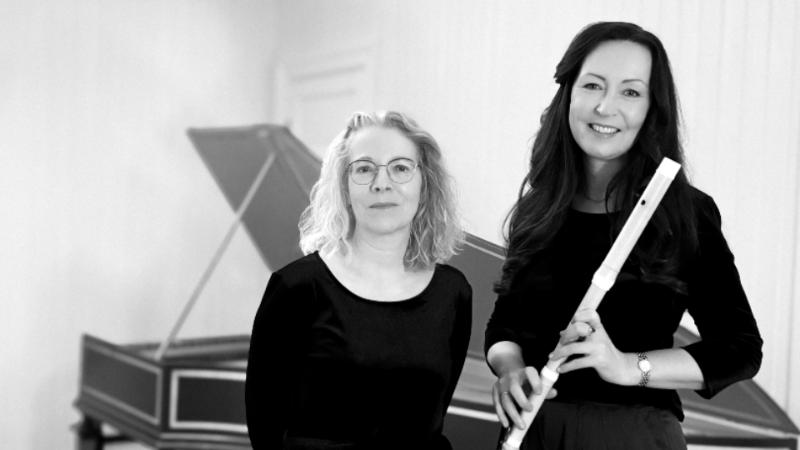 Two people standing beside each other in front of a harpsichord with one of them holding a baroque flute.